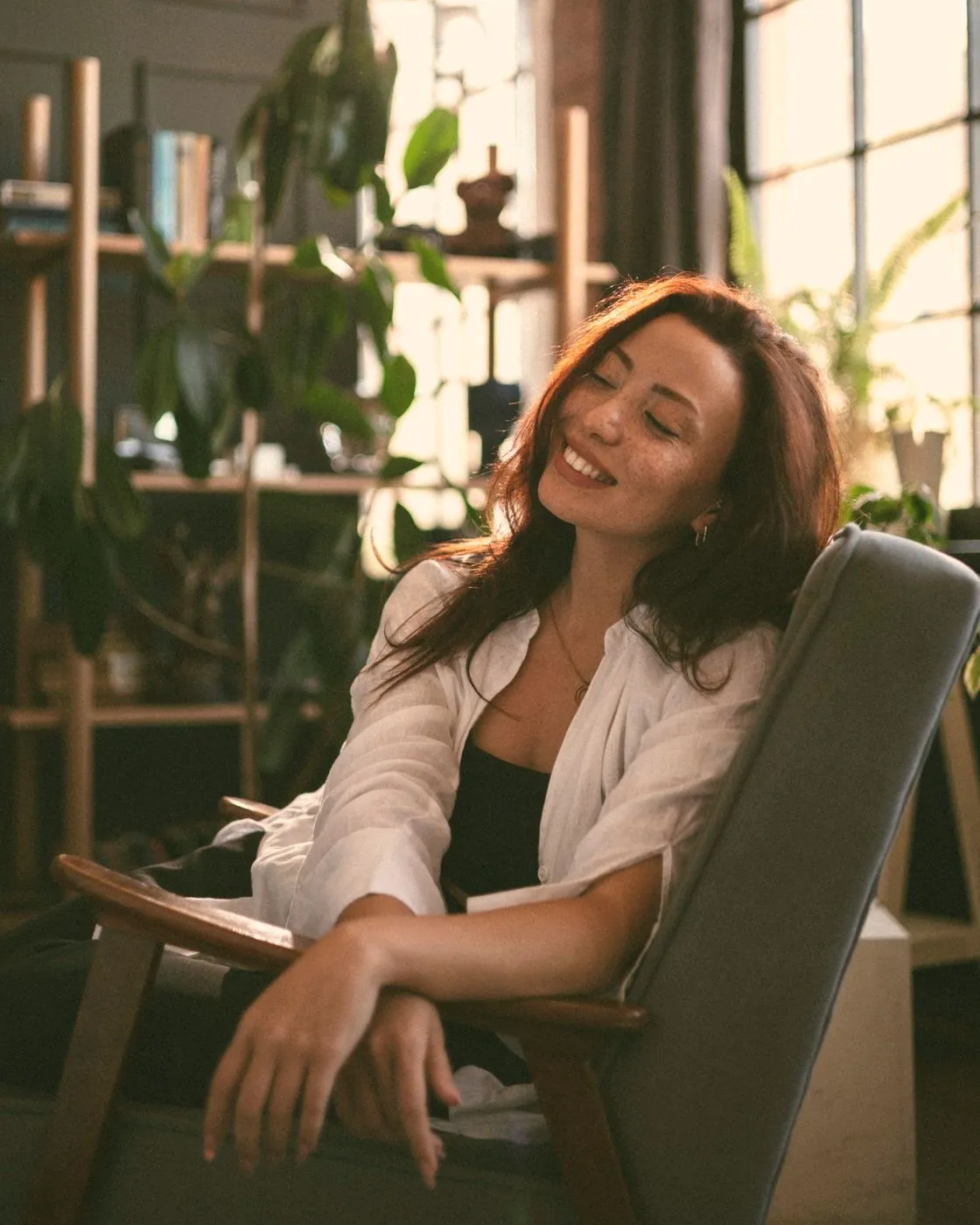 A woman leaning back in an armchair with her eyes closed and a smile, in a sunny room full of plants
