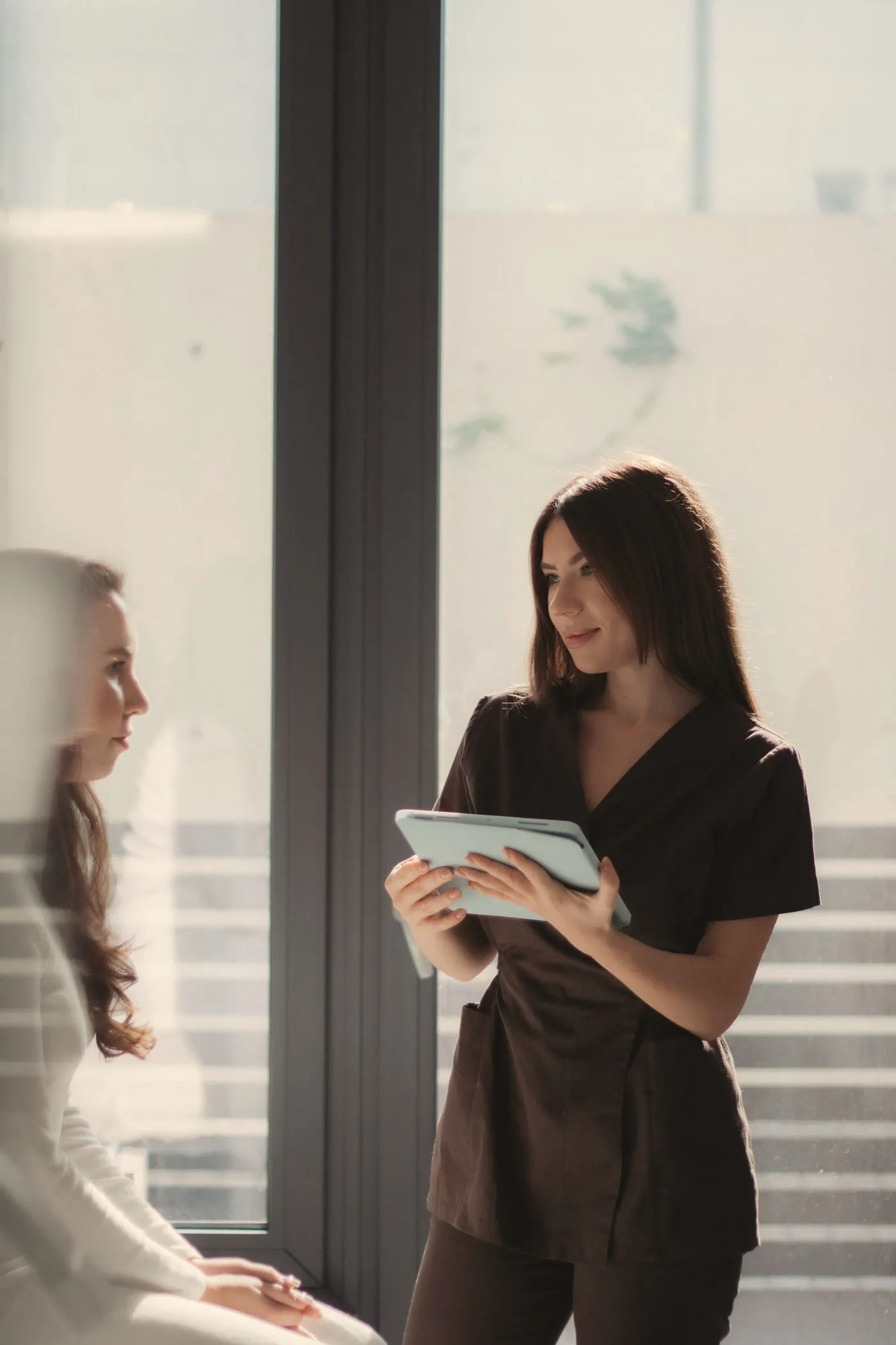 A clinician in dark scrubs holding a tablet and talking with a seated client beside a tall window