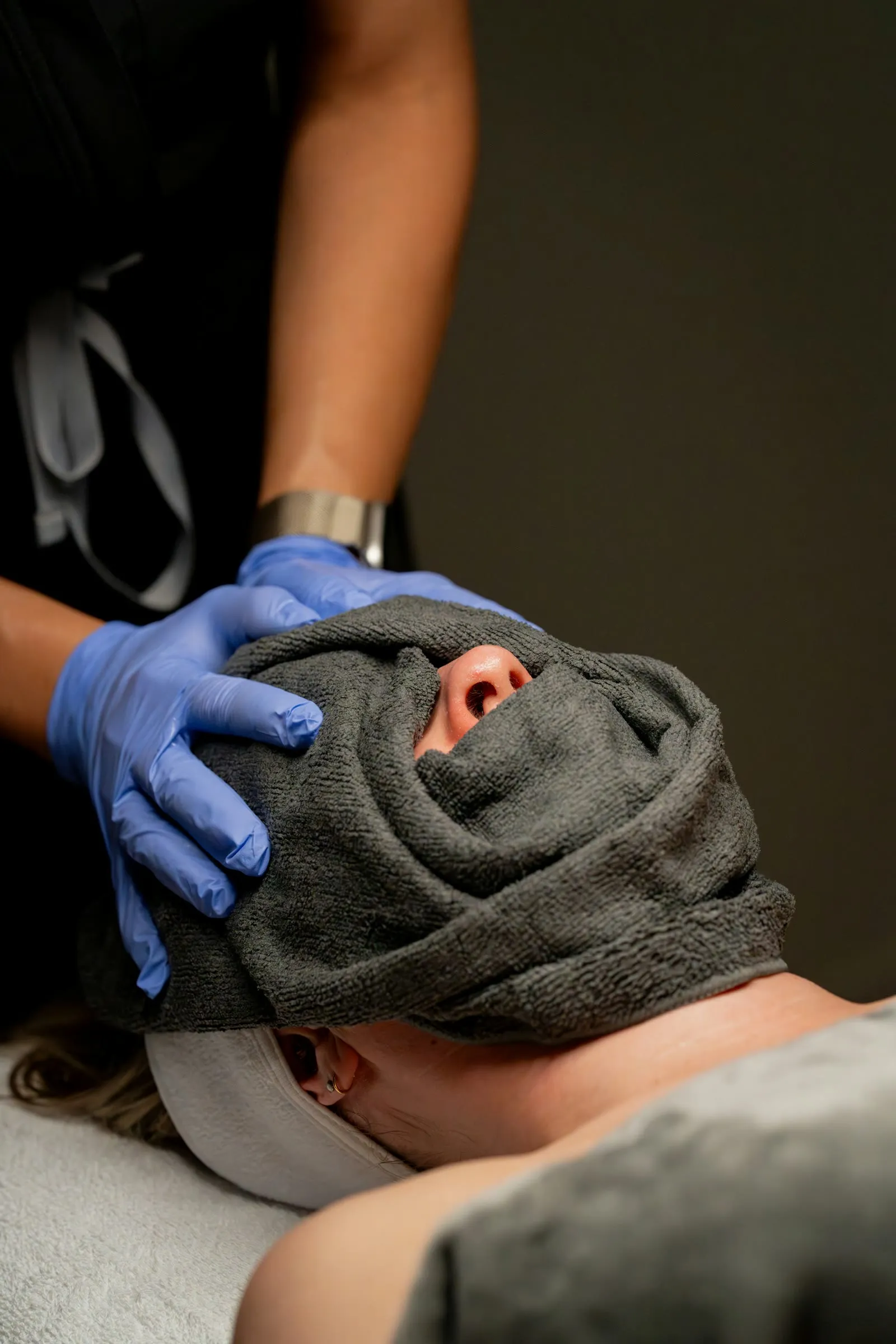 An esthetician in blue gloves wrapping a warm towel around a client lying on a treatment bed