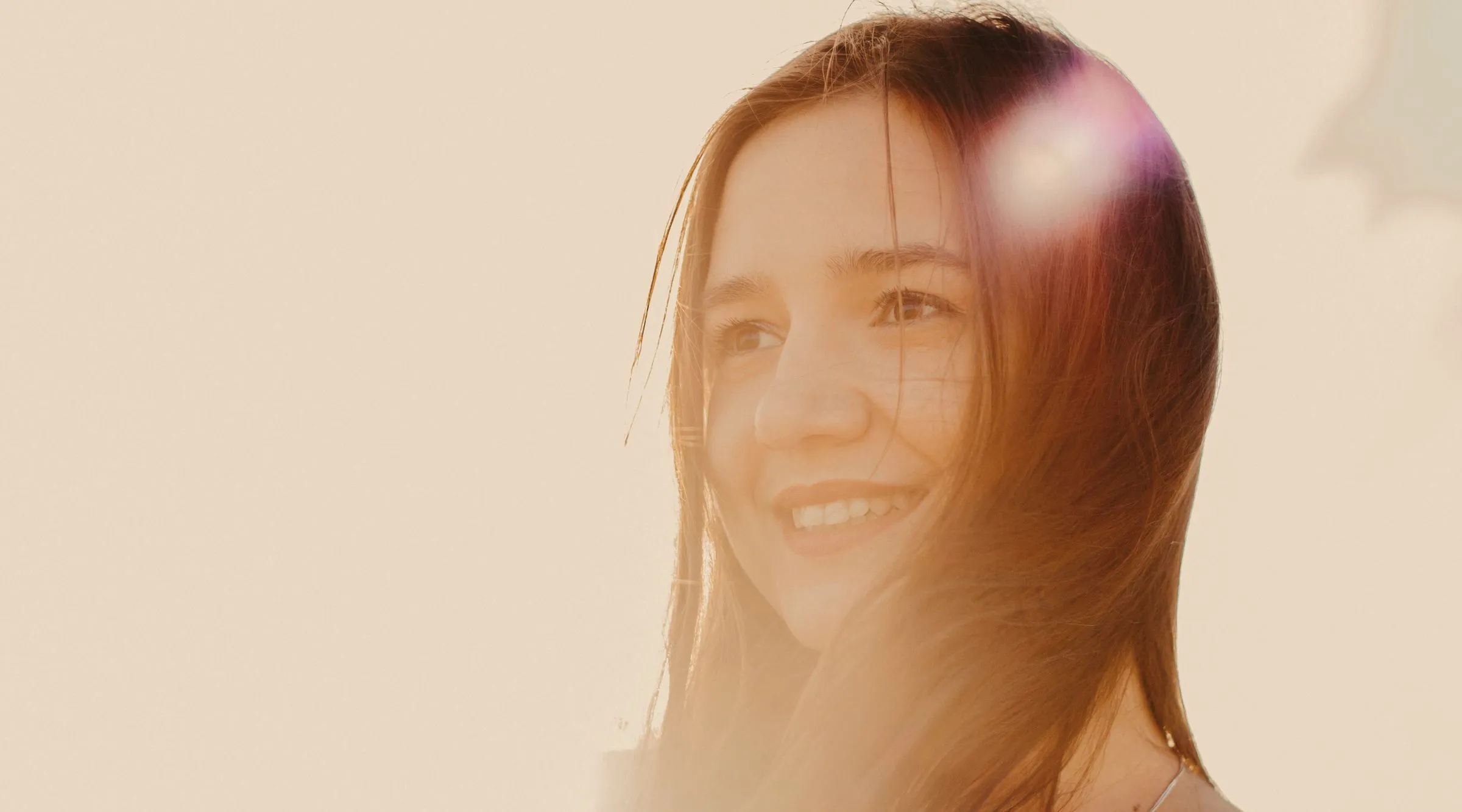 A young woman smiling in warm late-afternoon light, her hair lit gold against a pale cream sky