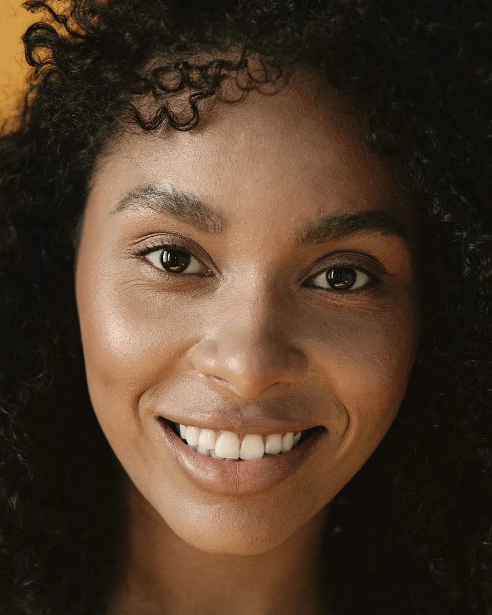 A woman with dark curly hair smiling at the camera against a warm yellow wall