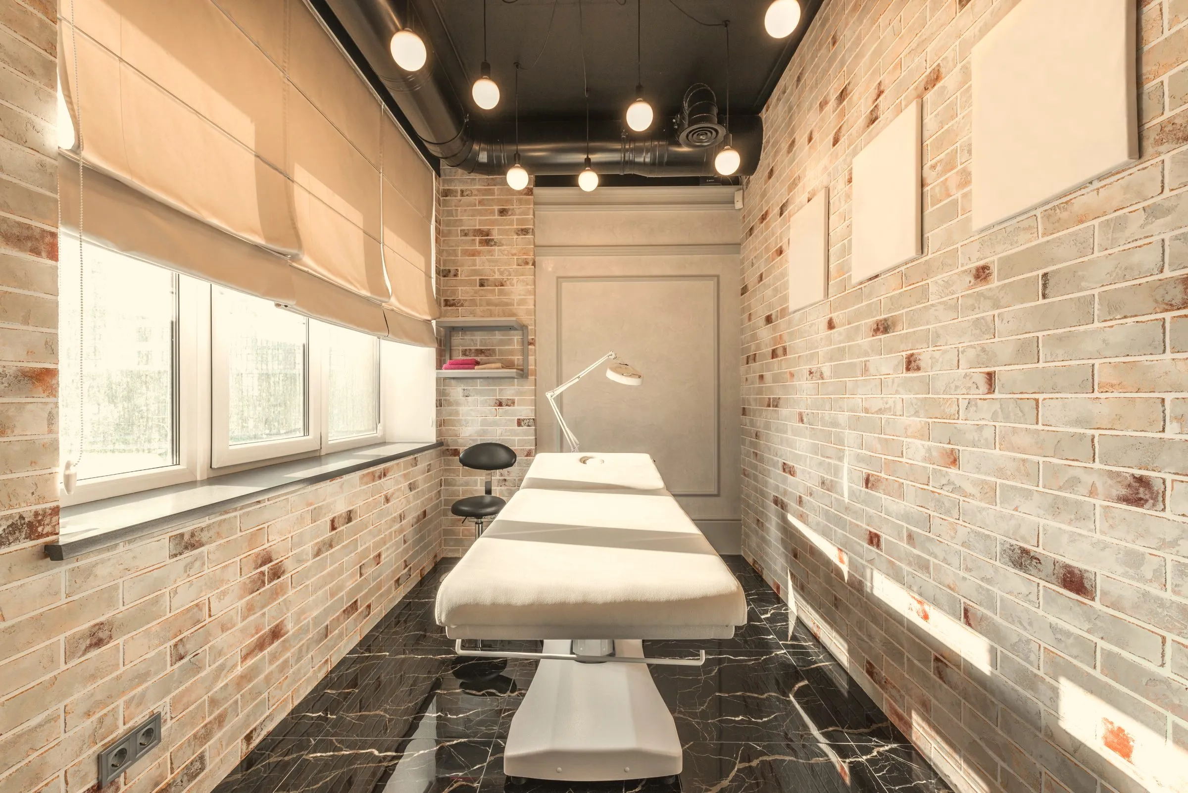 A made-up treatment bed in a narrow room with linen blinds, pale brick walls and daylight