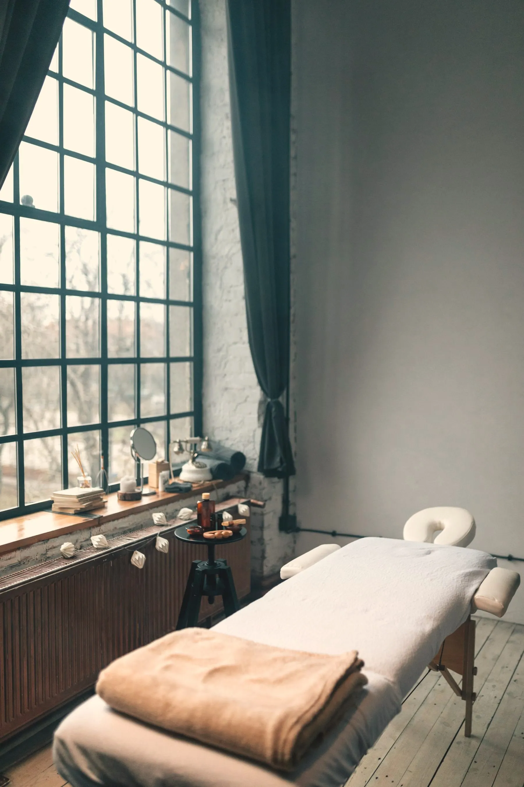 A treatment bed with a folded towel beside a tall window in daylight